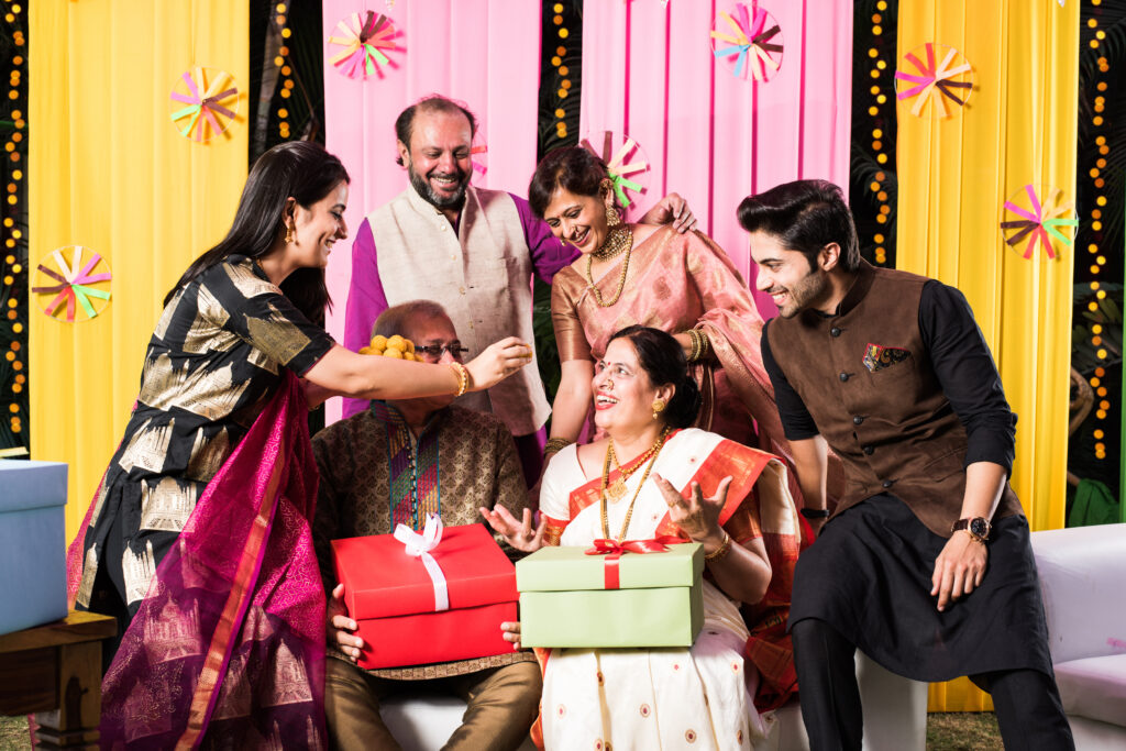 multigenerational indian family eating sweets while celebrating festival or occasion dressed in traditional wear, sitting on sofa or couch